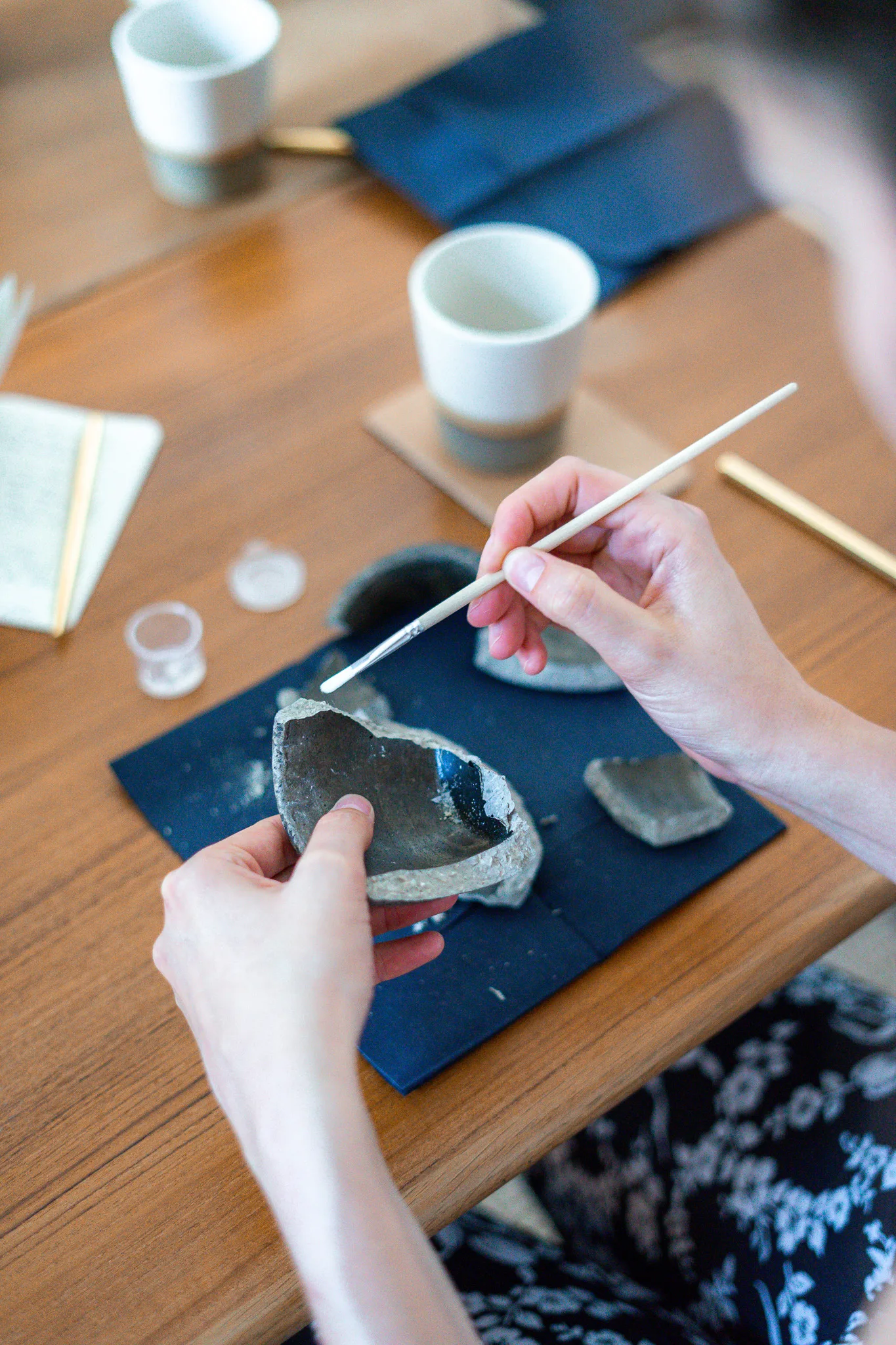 Zwei Hände halten eine zerbrochene Keramikschale und tragen mit einem feinen Pinsel Kintsugi-Kleber auf die Bruchkante auf